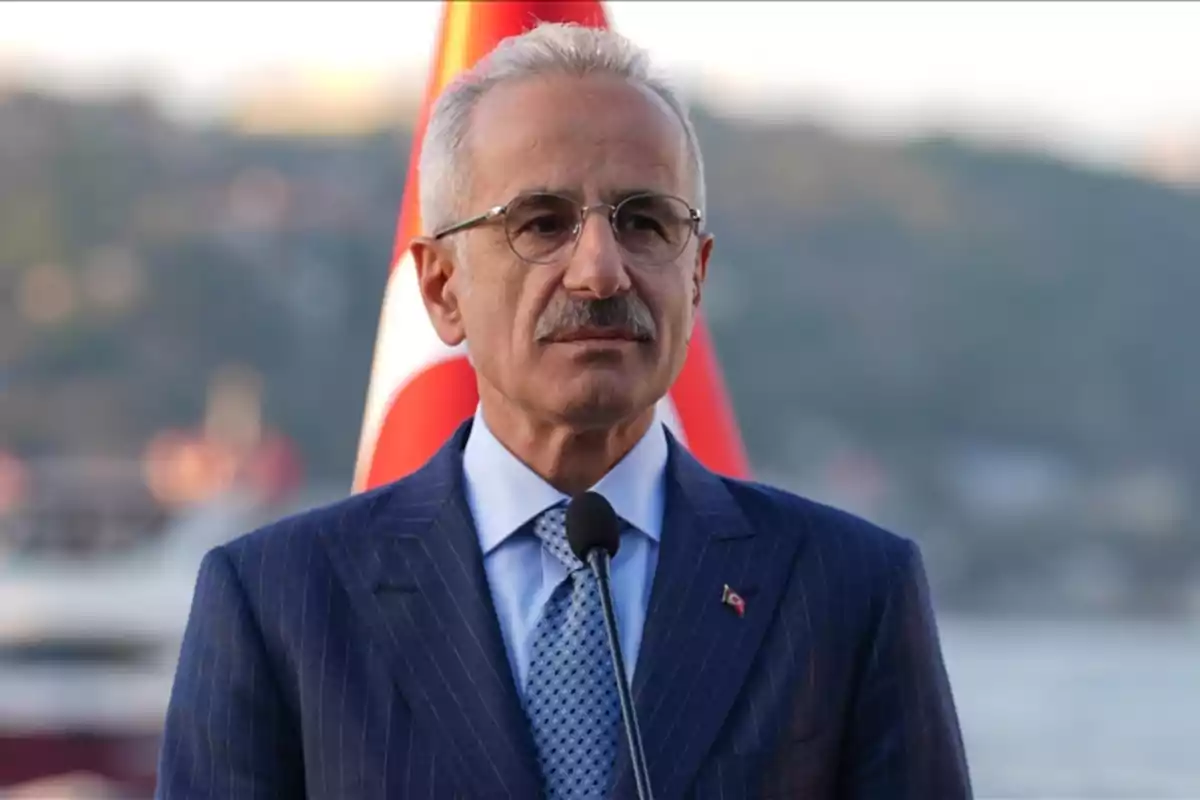 Gray-haired man with glasses wearing a blue suit and tie, standing in front of a microphone with a blurred Turkish flag in the background Gray-haired man with glasses wearing a blue suit and tie, standing in front of a microphone with a blurred Turkish flag in the background