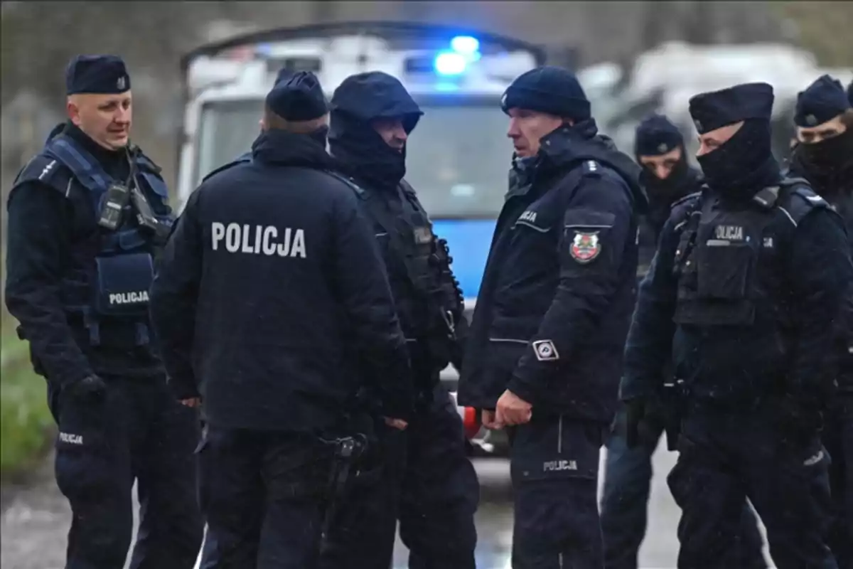 Several police officers in dark uniforms and bulletproof vests are gathered in the street in front of a patrol car with its lights on. Several police officers in dark uniforms and bulletproof vests are gathered in the street in front of a patrol car with its lights on.