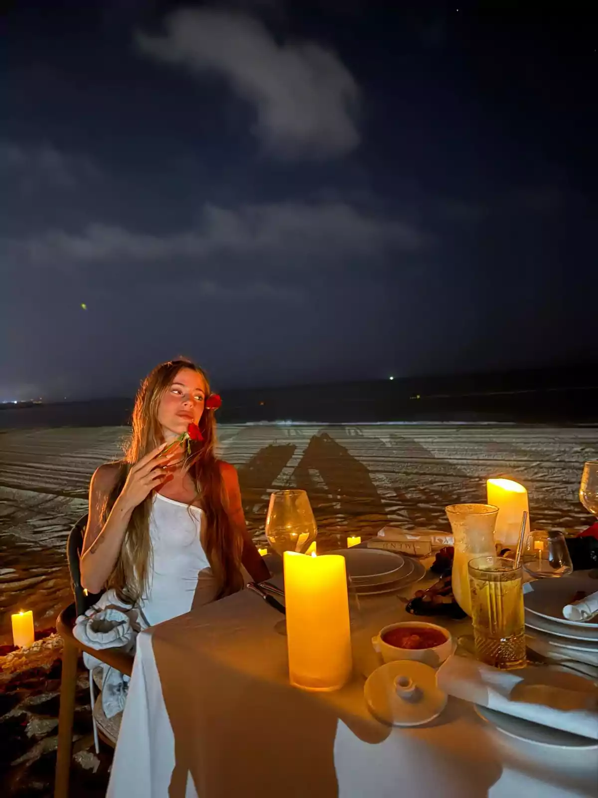 Woman sitting at an elegantly decorated table with candles on the beach at night, holding a red flower and looking to the side Woman sitting at an elegantly decorated table with candles on the beach at night, holding a red flower and looking to the side