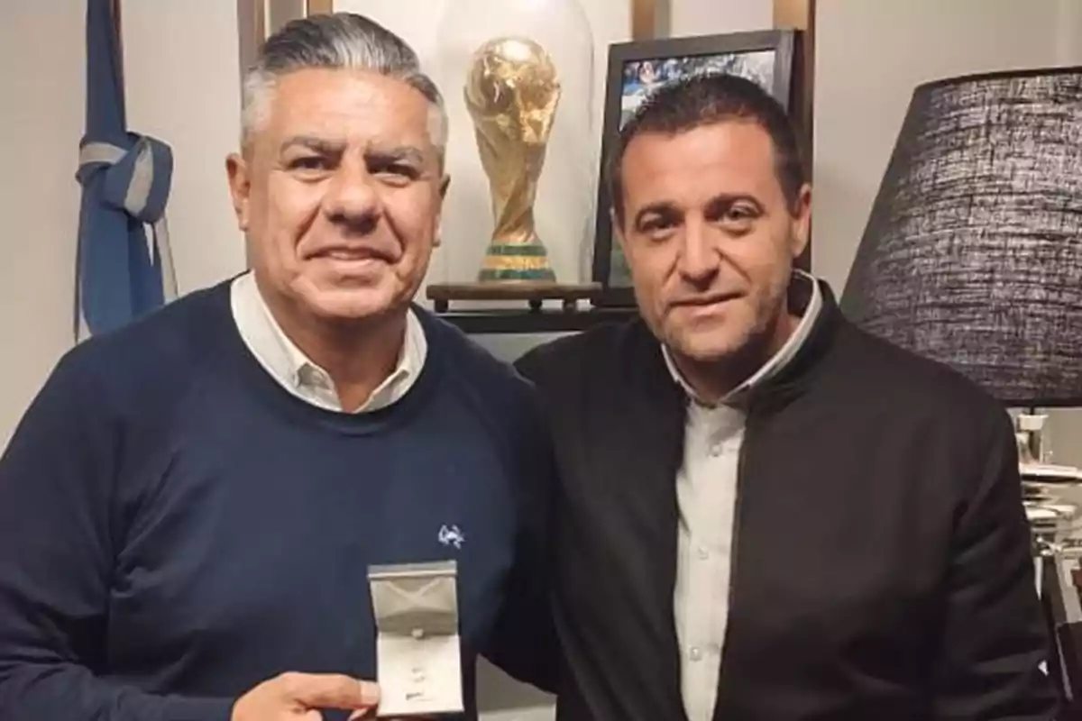 Two men pose together in an office, one of them holding a box with a medal, and behind them there is a replica of the World Cup trophy. Two men pose together in an office, one of them holding a box with a medal, and behind them there is a replica of the World Cup trophy.