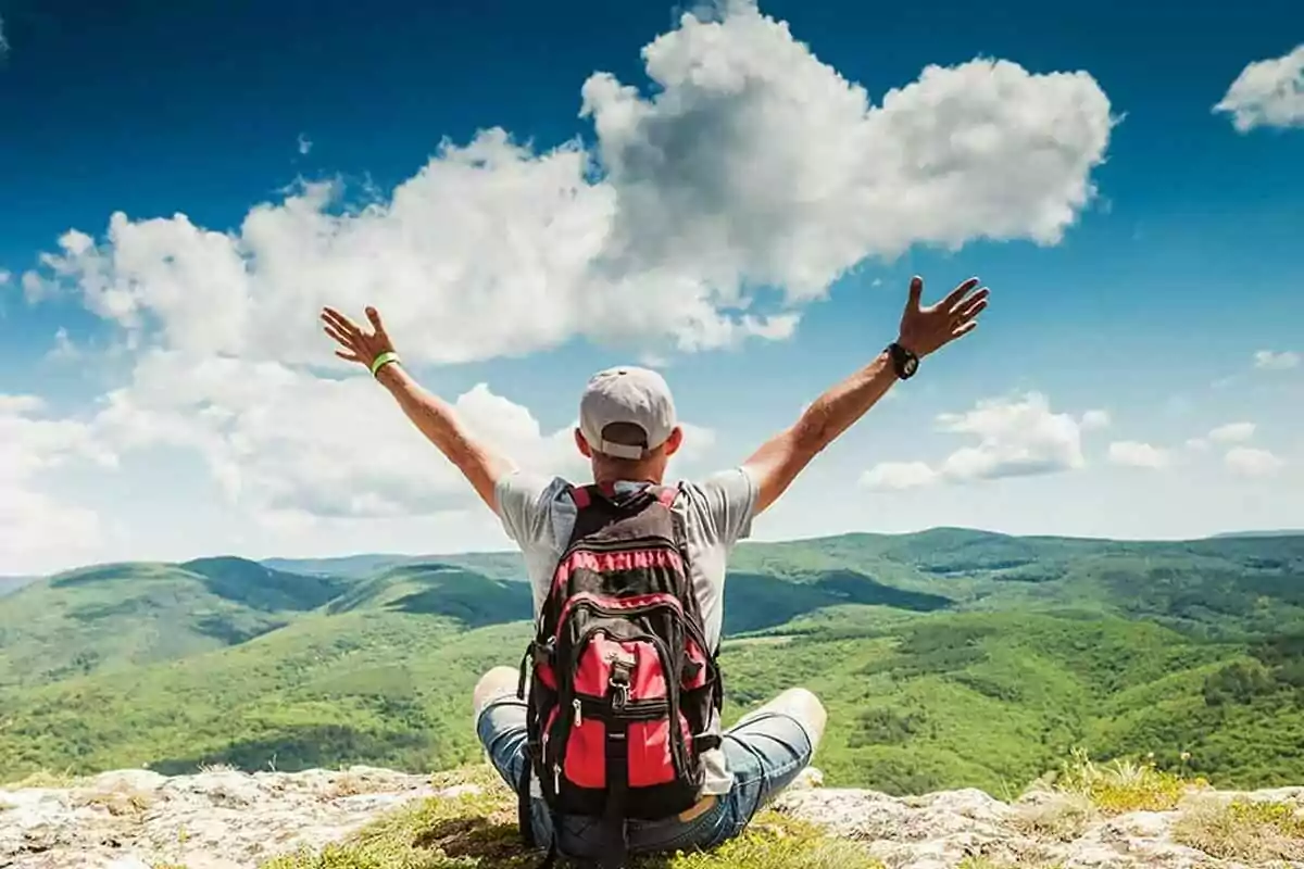 Persona sentada en la cima de una montaña con los brazos extendidos disfrutando de la vista panorámica de un paisaje verde bajo un cielo azul con nubes Persona sentada en la cima de una montaña con los brazos extendidos disfrutando de la vista panorámica de un paisaje verde bajo un cielo azul con nubes