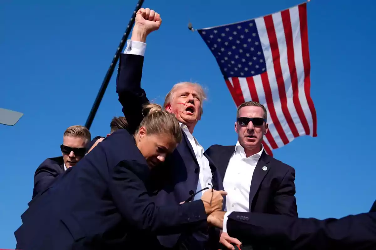 A man with an injured face raises his fist while being protected by several people in dark suits and sunglasses; in the background, an American flag waves under a blue sky. A man with an injured face raises his fist while being protected by several people in dark suits and sunglasses; in the background, an American flag waves under a blue sky.