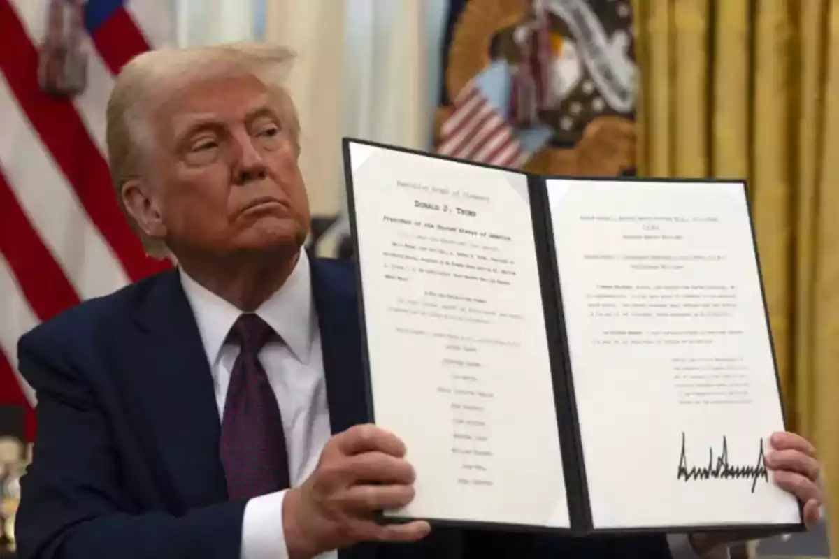 Hombre de cabello rubio con traje oscuro sostiene un documento abierto con texto y una firma visible en una oficina con banderas al fondo Hombre de cabello rubio con traje oscuro sostiene un documento abierto con texto y una firma visible en una oficina con banderas al fondo