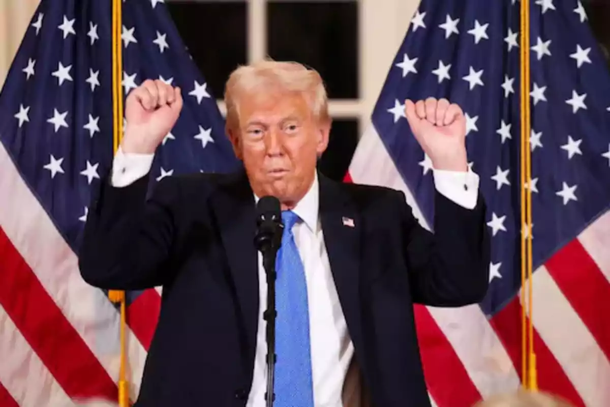 Hombre de cabello rubio con traje oscuro y corbata azul levantando ambos brazos frente a dos banderas de Estados Unidos Hombre de cabello rubio con traje oscuro y corbata azul levantando ambos brazos frente a dos banderas de Estados Unidos