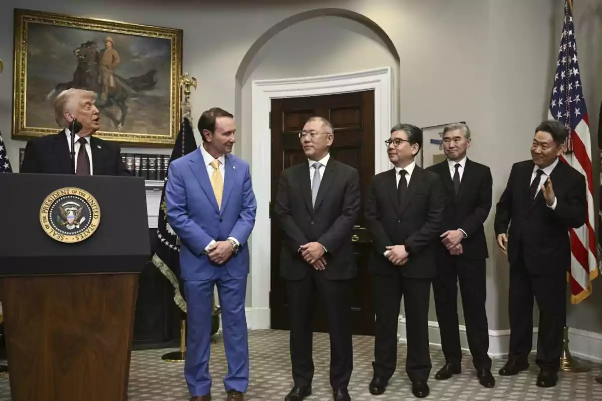 Un grupo de seis hombres con trajes formales está reunido en una sala oficial con banderas de Estados Unidos y un podio presidencial, mientras uno de ellos habla y los demás lo observan.