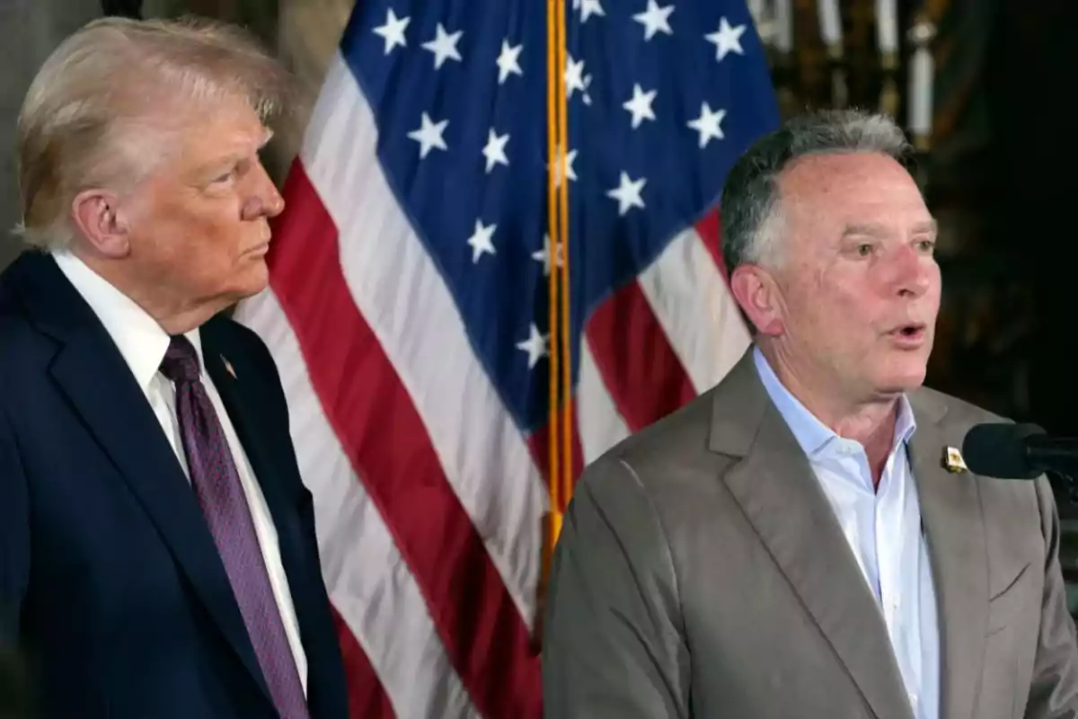 Two men standing in front of a United States flag while one of them speaks into a microphone Two men standing in front of a United States flag while one of them speaks into a microphone