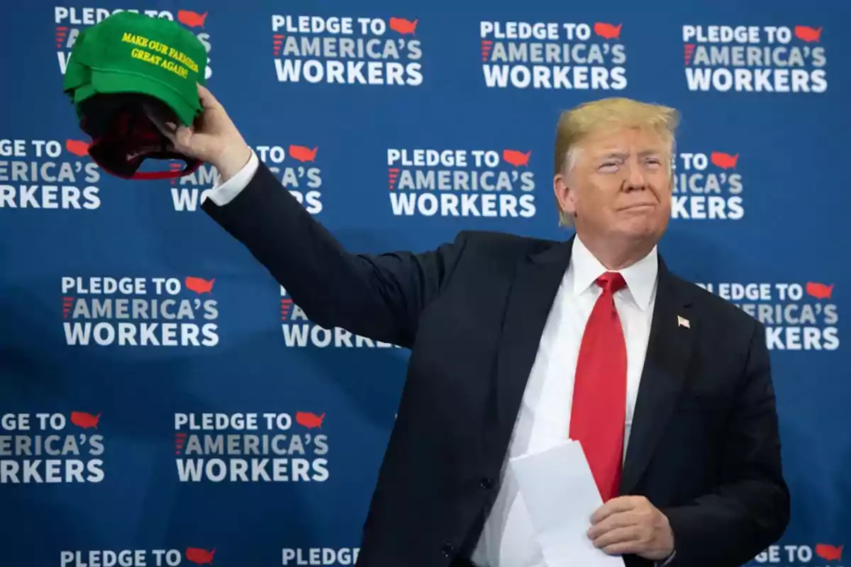 Man in a dark suit and red tie holds several green caps with the phrase Make Our Farmers Great Again in front of a blue background with the text Pledge to America's Workers. Man in a dark suit and red tie holds several green caps with the phrase Make Our Farmers Great Again in front of a blue background with the text Pledge to America's Workers.