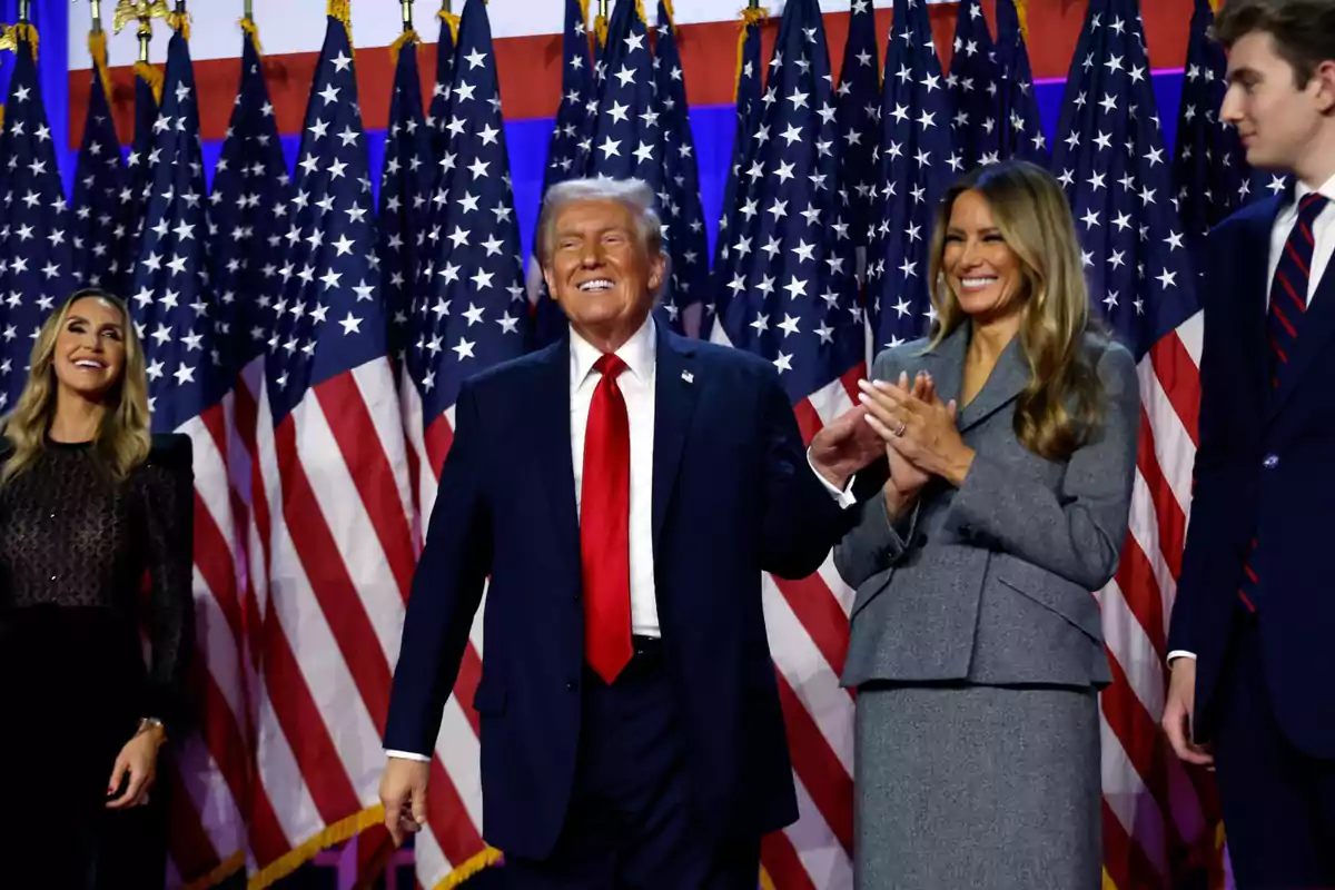 A man in a dark suit and red tie smiles while accompanied by three people in front of several American flags. A man in a dark suit and red tie smiles while accompanied by three people in front of several American flags.