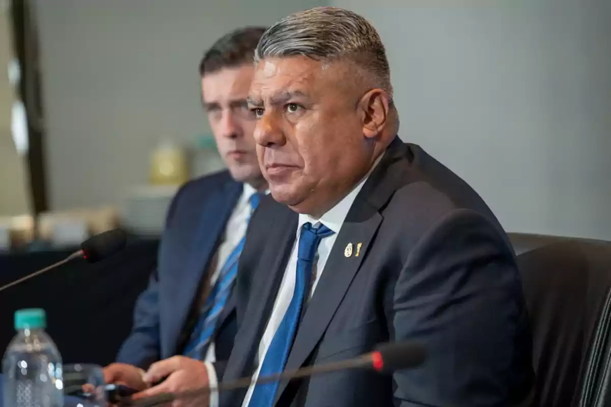 Dos hombres con traje y corbata sentados en una mesa de conferencia frente a micrófonos Dos hombres con traje y corbata sentados en una mesa de conferencia frente a micrófonos