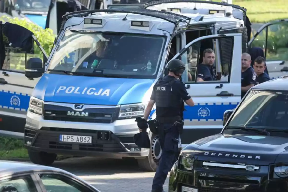 Several police officers in uniform and bulletproof vests are standing next to a blue and white police van with the word "Policja" on the front, while one of them walks toward the vehicle and others watch from inside.