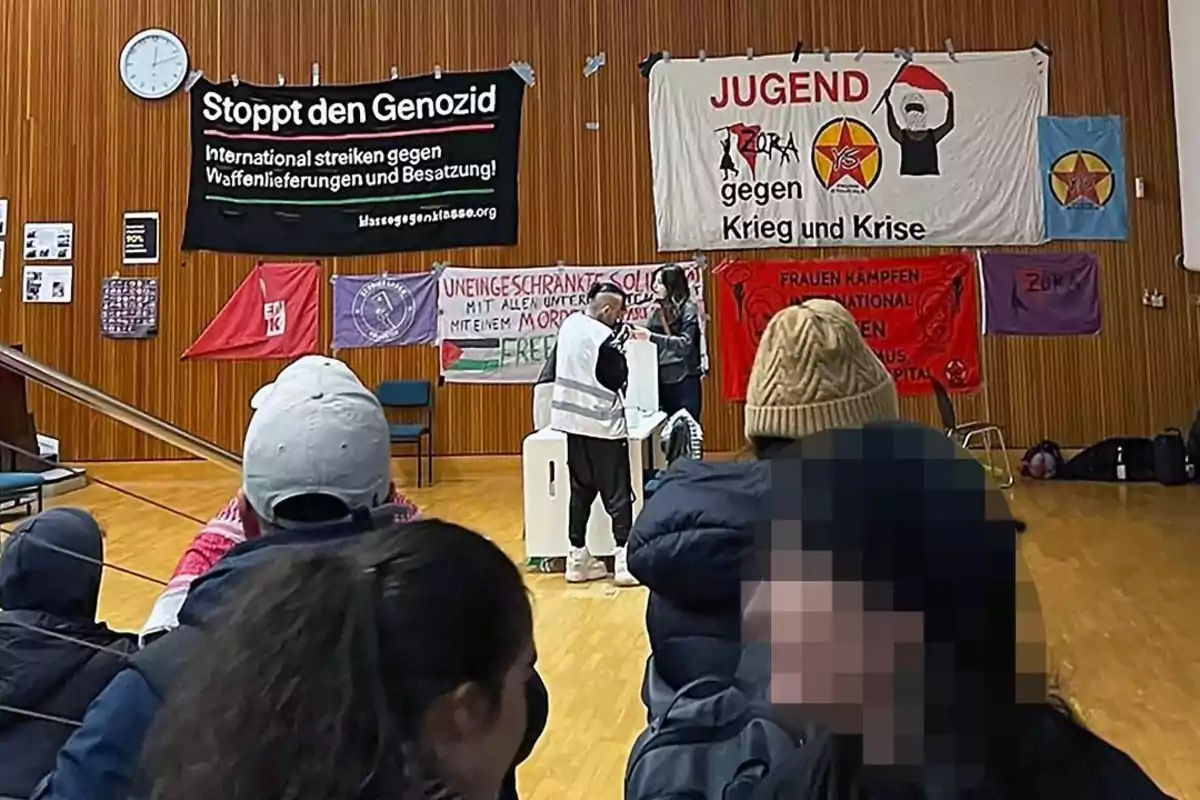 Personas sentadas observan a un orador en una sala con pancartas de protesta en las paredes, algunas con mensajes en alemán sobre genocidio, guerra y crisis. Personas sentadas observan a un orador en una sala con pancartas de protesta en las paredes, algunas con mensajes en alemán sobre genocidio, guerra y crisis.