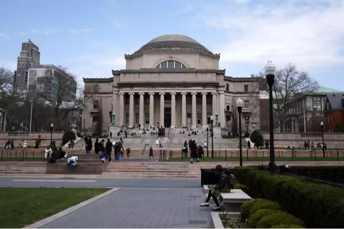 Edificio universitario de estilo clásico con columnas y cúpula, rodeado de personas sentadas y caminando en un campus al aire libre