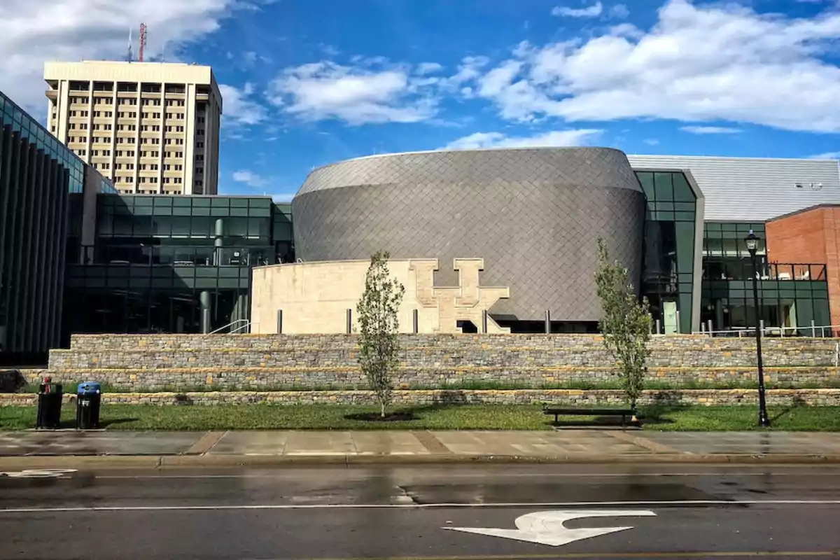 Modern building with a glass facade and circular metal structure, featuring a large letter K logo at the front and a blue sky with clouds in the background