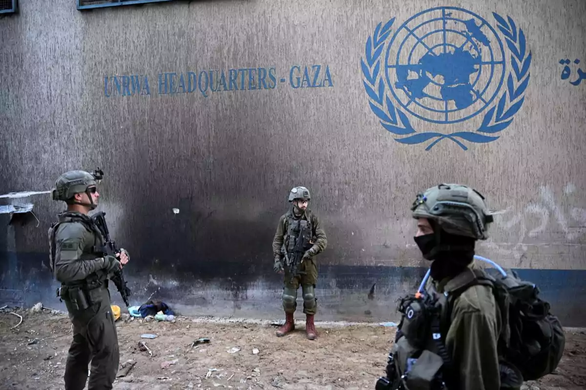 Tres soldados armados con uniforme militar y equipo táctico están frente a un edificio con el logo de la ONU y la inscripción UNRWA Headquarters Gaza en la pared.