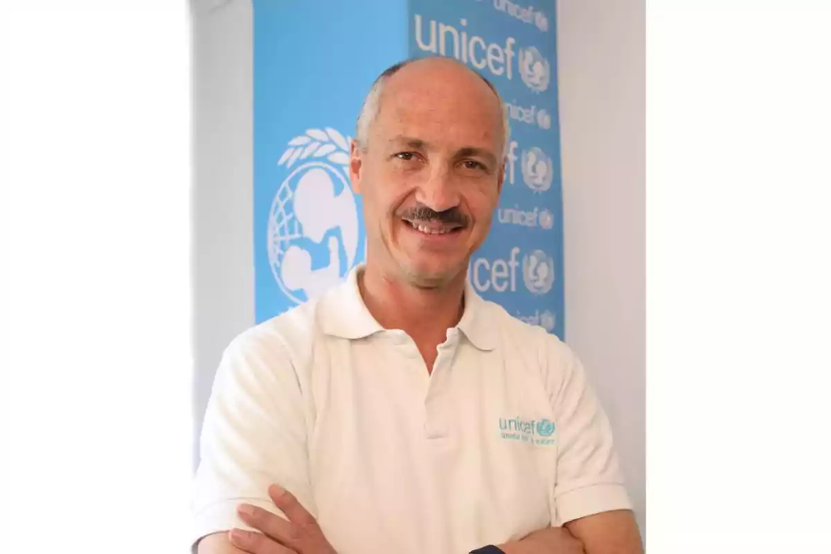 Hombre de mediana edad con bigote y camiseta blanca de Unicef sonriendo frente a un cartel azul de Unicef