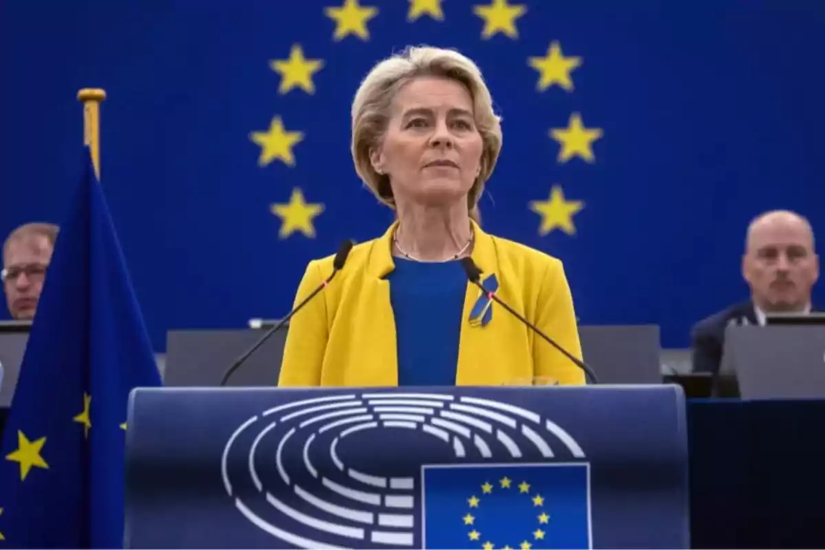Blonde-haired woman wearing a yellow jacket and blue blouse speaking at a podium in front of a background with yellow stars on blue representing the flag of the European Union Blonde-haired woman wearing a yellow jacket and blue blouse speaking at a podium in front of a background with yellow stars on blue representing the flag of the European Union