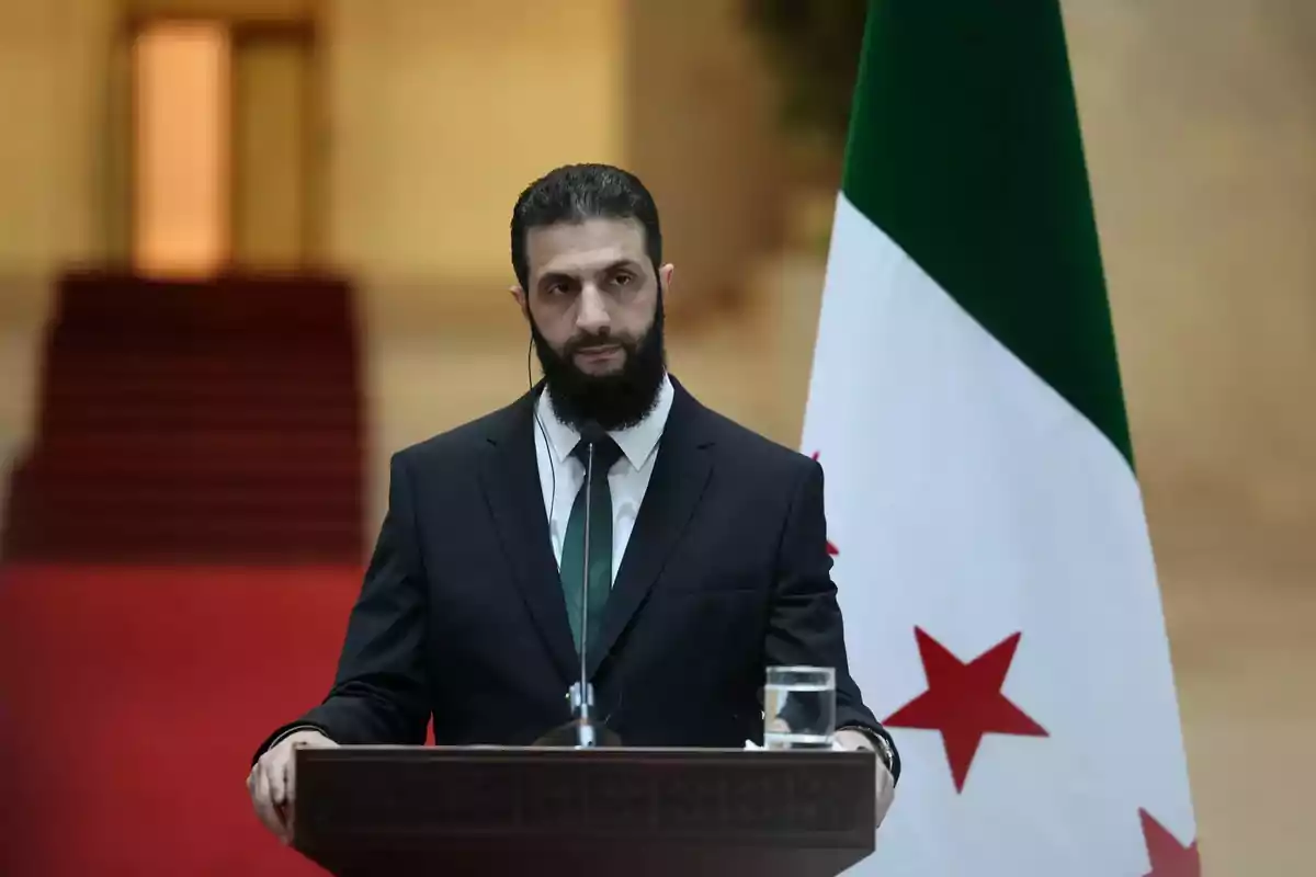 Hombre de barba oscura con traje y corbata hablando en un podio junto a una bandera con estrellas rojas y fondo desenfocado Hombre de barba oscura con traje y corbata hablando en un podio junto a una bandera con estrellas rojas y fondo desenfocado
