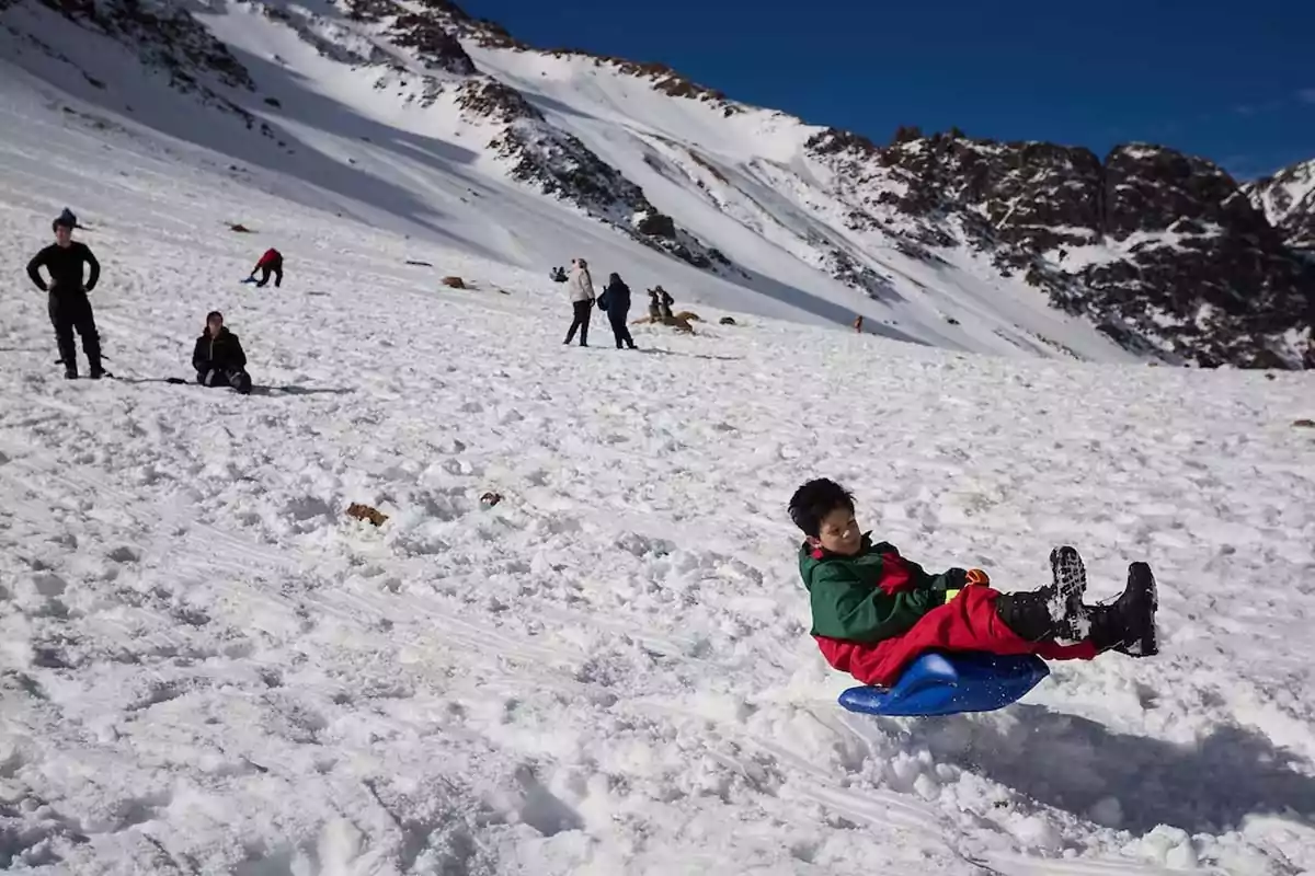 Child sliding in the snow on a blue sled while other people enjoy the snowy landscape and mountains in the background Child sliding in the snow on a blue sled while other people enjoy the snowy landscape and mountains in the background