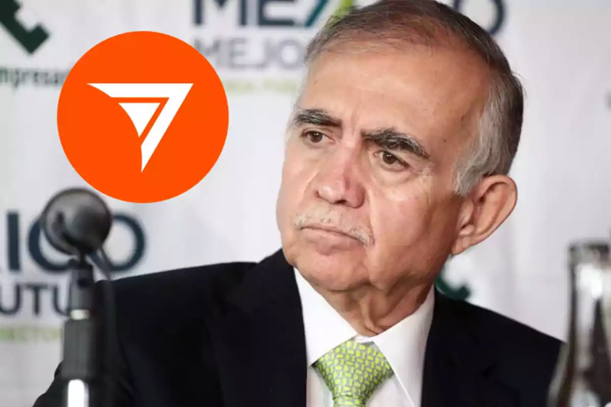 Hombre de cabello canoso y bigote con expresión seria, usando traje y corbata verde, sentado frente a un micrófono con un fondo de logotipos y un círculo naranja con un símbolo blanco