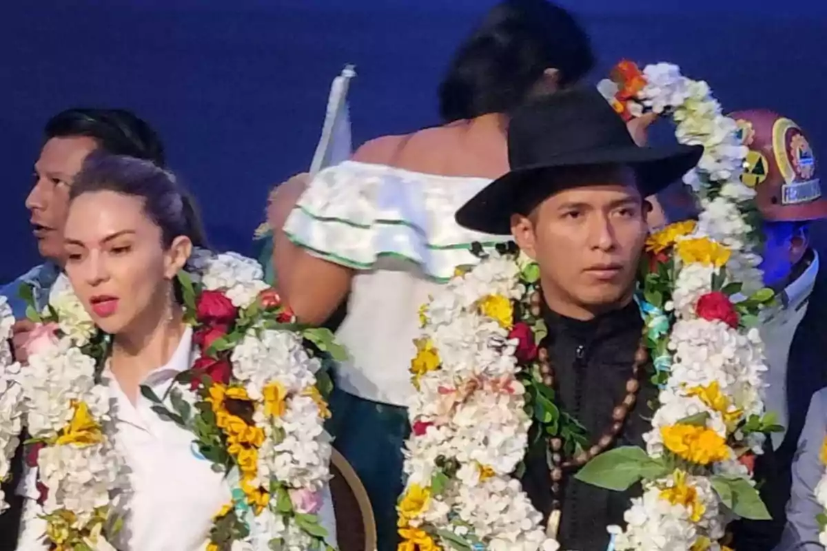 Dos personas con collares de flores y ropa tradicional en un evento, una de ellas lleva sombrero negro y la otra viste de blanco.