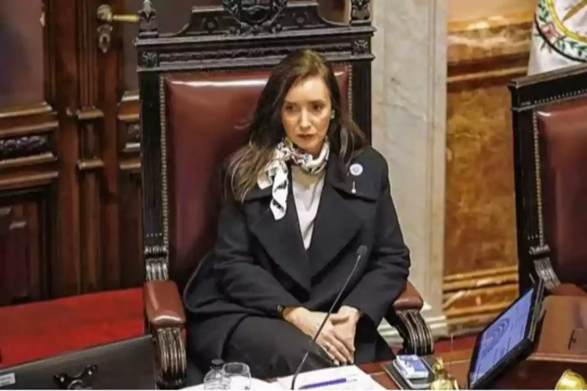 Mujer sentada en una silla grande de madera en un recinto parlamentario con expresión seria y vestida con abrigo oscuro y pañuelo al cuello Mujer sentada en una silla grande de madera en un recinto parlamentario con expresión seria y vestida con abrigo oscuro y pañuelo al cuello