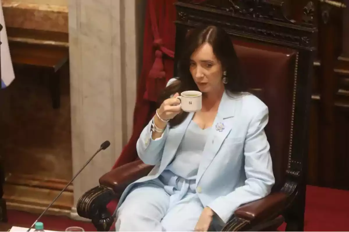 Mujer sentada en una silla grande de madera, vestida con un traje celeste claro, sosteniendo una taza blanca y bebiendo en un ambiente formal