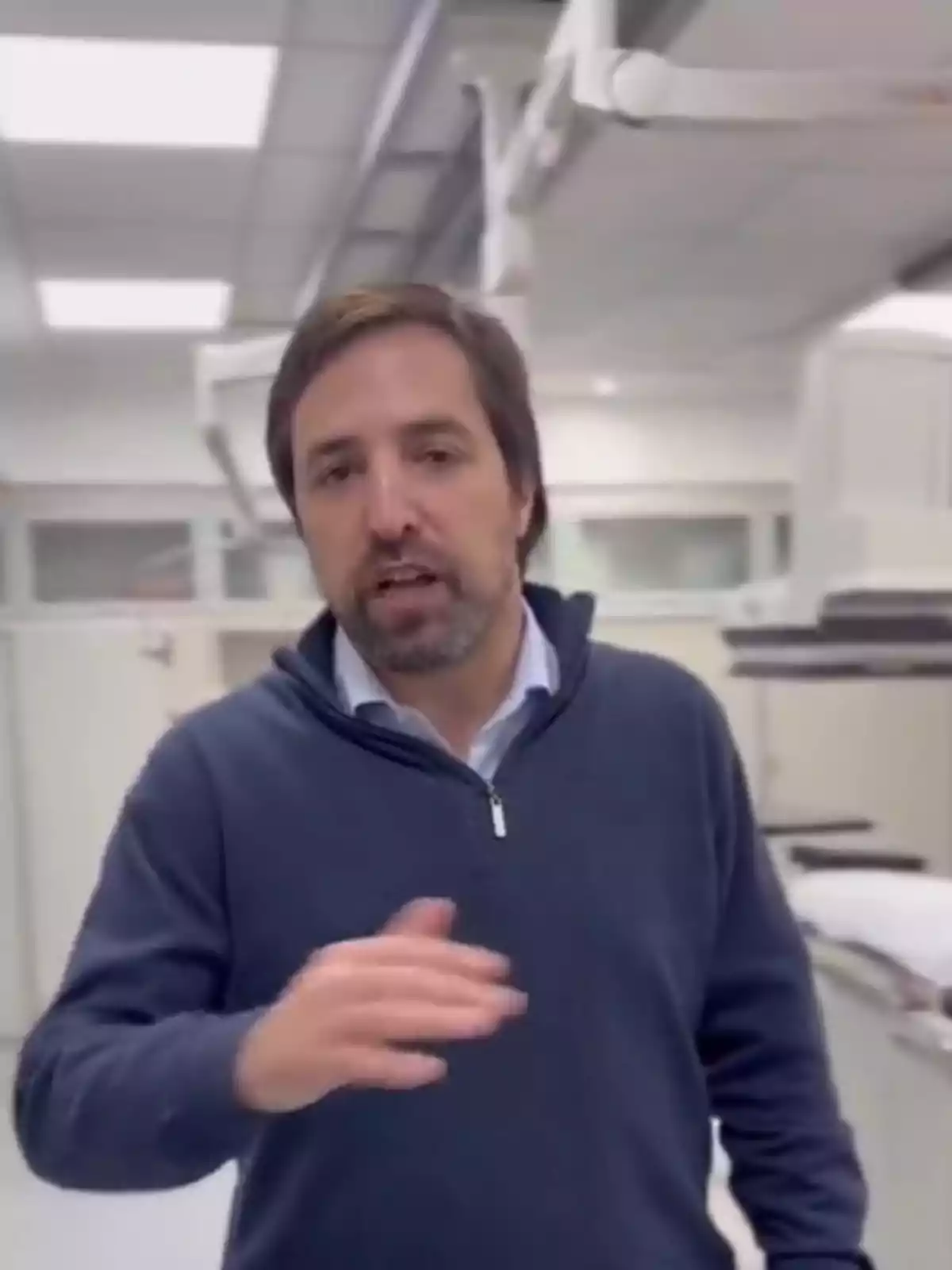 Hombre de cabello castaño y barba corta con suéter azul marino y camisa clara, hablando y gesticulando en lo que parece ser una sala de hospital o laboratorio clínico. Hombre de cabello castaño y barba corta con suéter azul marino y camisa clara, hablando y gesticulando en lo que parece ser una sala de hospital o laboratorio clínico.