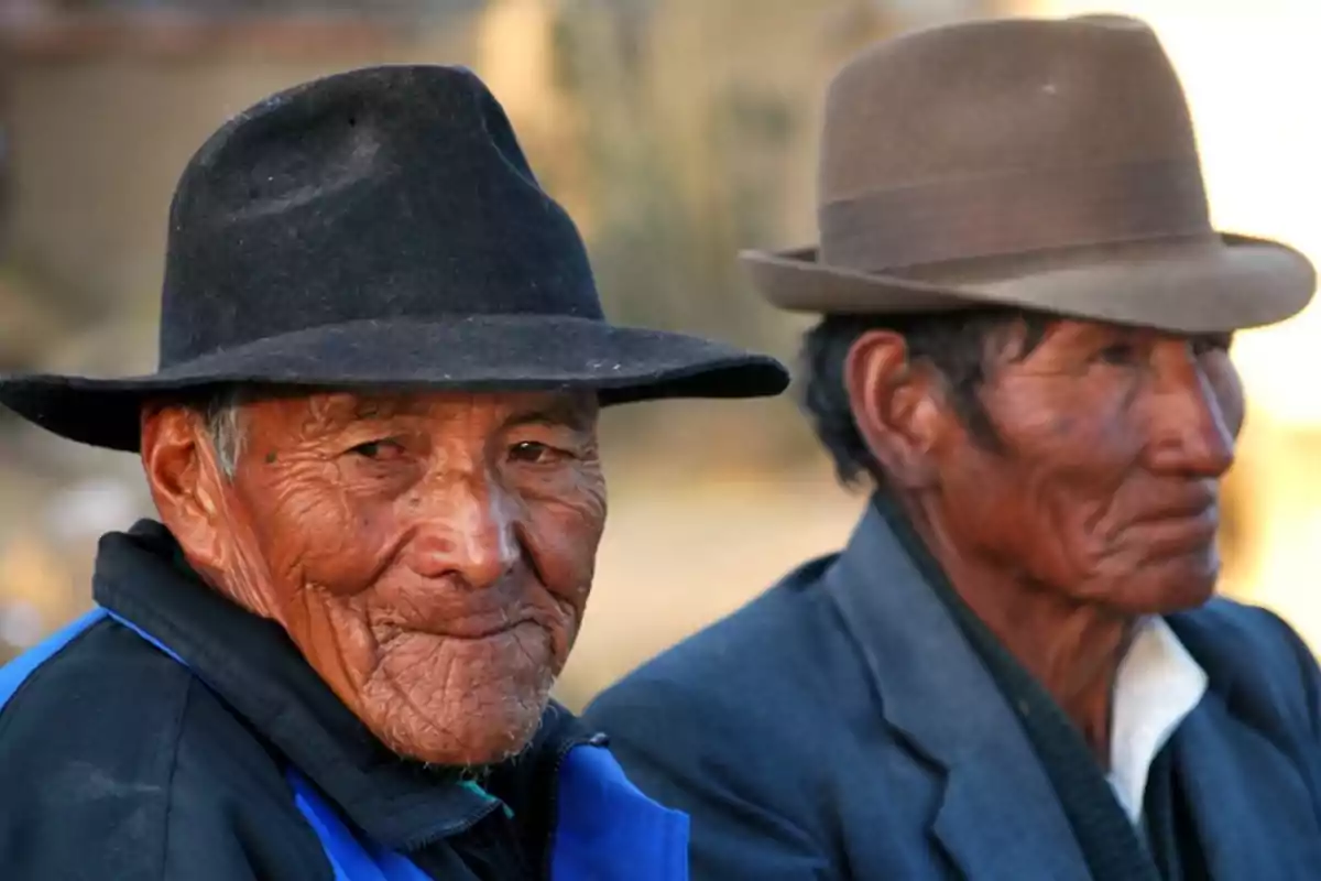 Dos hombres mayores con sombreros tradicionales y rostros arrugados miran hacia la cámara en un entorno exterior iluminado por la luz del sol