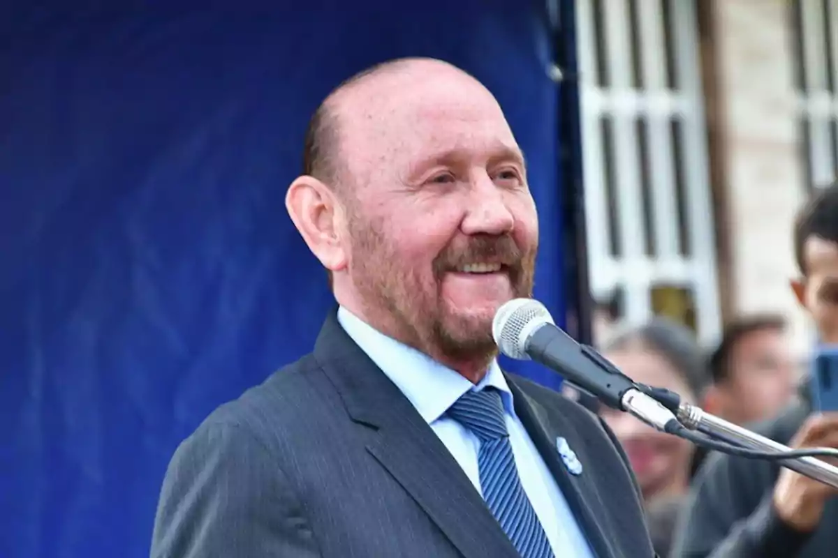 Hombre de traje y corbata azul sonriendo mientras habla frente a un micrófono en un evento al aire libre