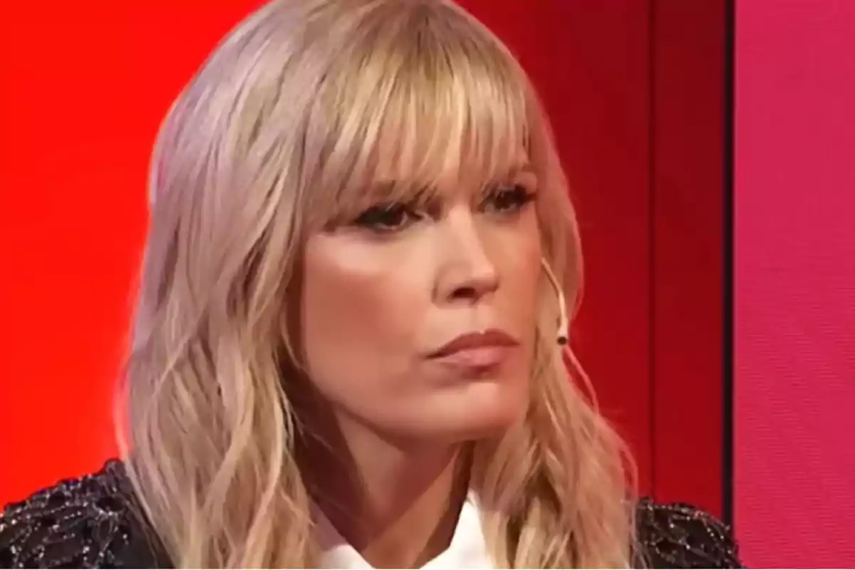 Mujer rubia con flequillo y expresión seria en un set de televisión con fondo rojo Mujer rubia con flequillo y expresión seria en un set de televisión con fondo rojo