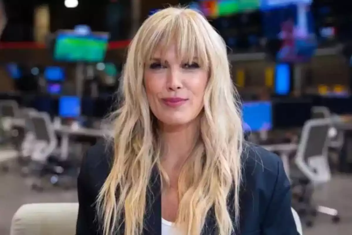 Mujer rubia de cabello largo y lacio con flequillo, vestida con saco oscuro, sentada en una oficina moderna con varias pantallas y escritorios al fondo