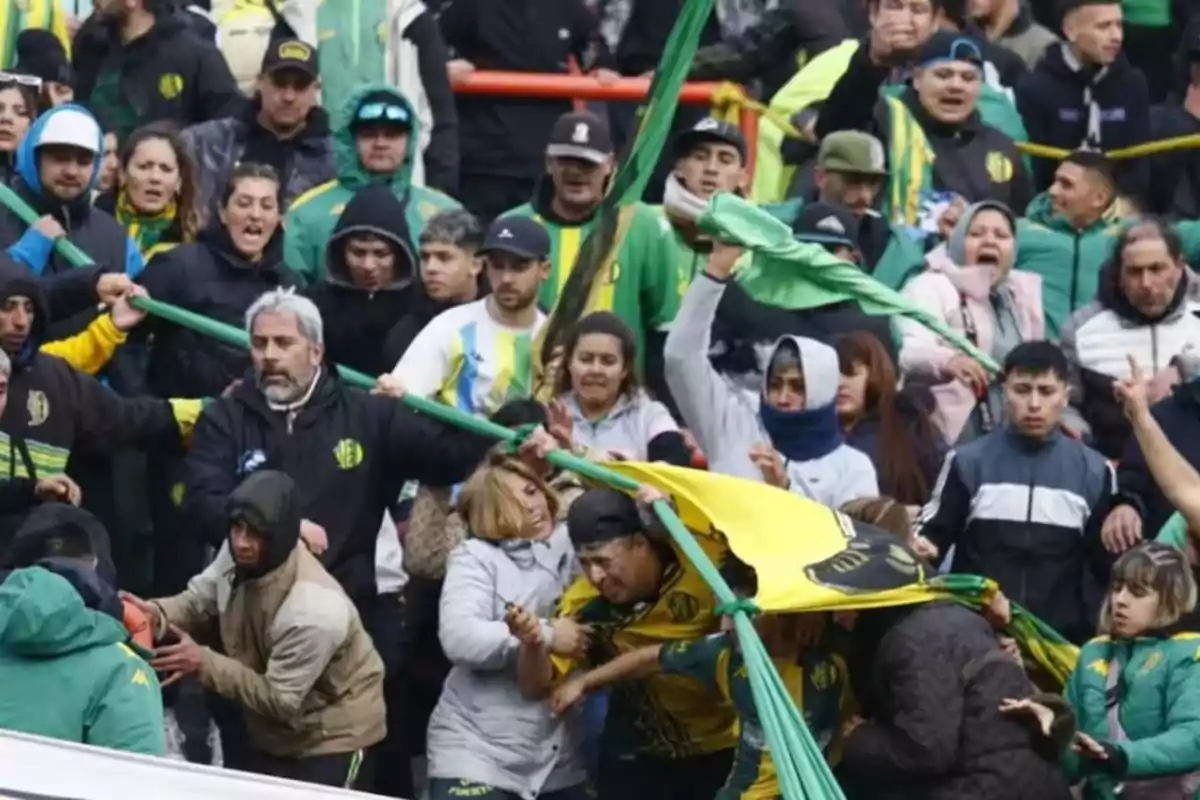 Aficionados de fútbol discutiendo y empujándose en las gradas durante un partido, algunos con banderas y bufandas del equipo, mostrando tensión y desorden.