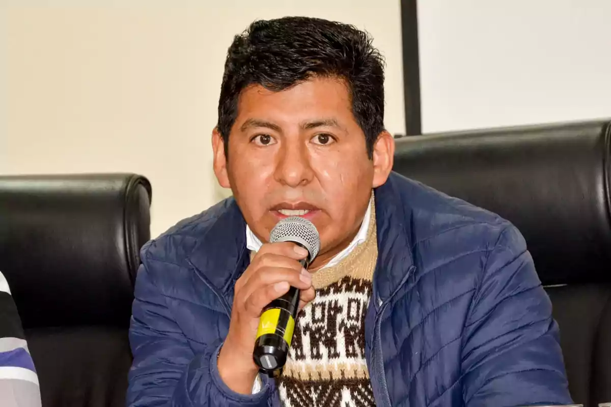 Hombre de cabello corto y oscuro hablando por un micrófono mientras está sentado en una silla negra, usando un suéter con diseño andino y una chaqueta azul Hombre de cabello corto y oscuro hablando por un micrófono mientras está sentado en una silla negra, usando un suéter con diseño andino y una chaqueta azul