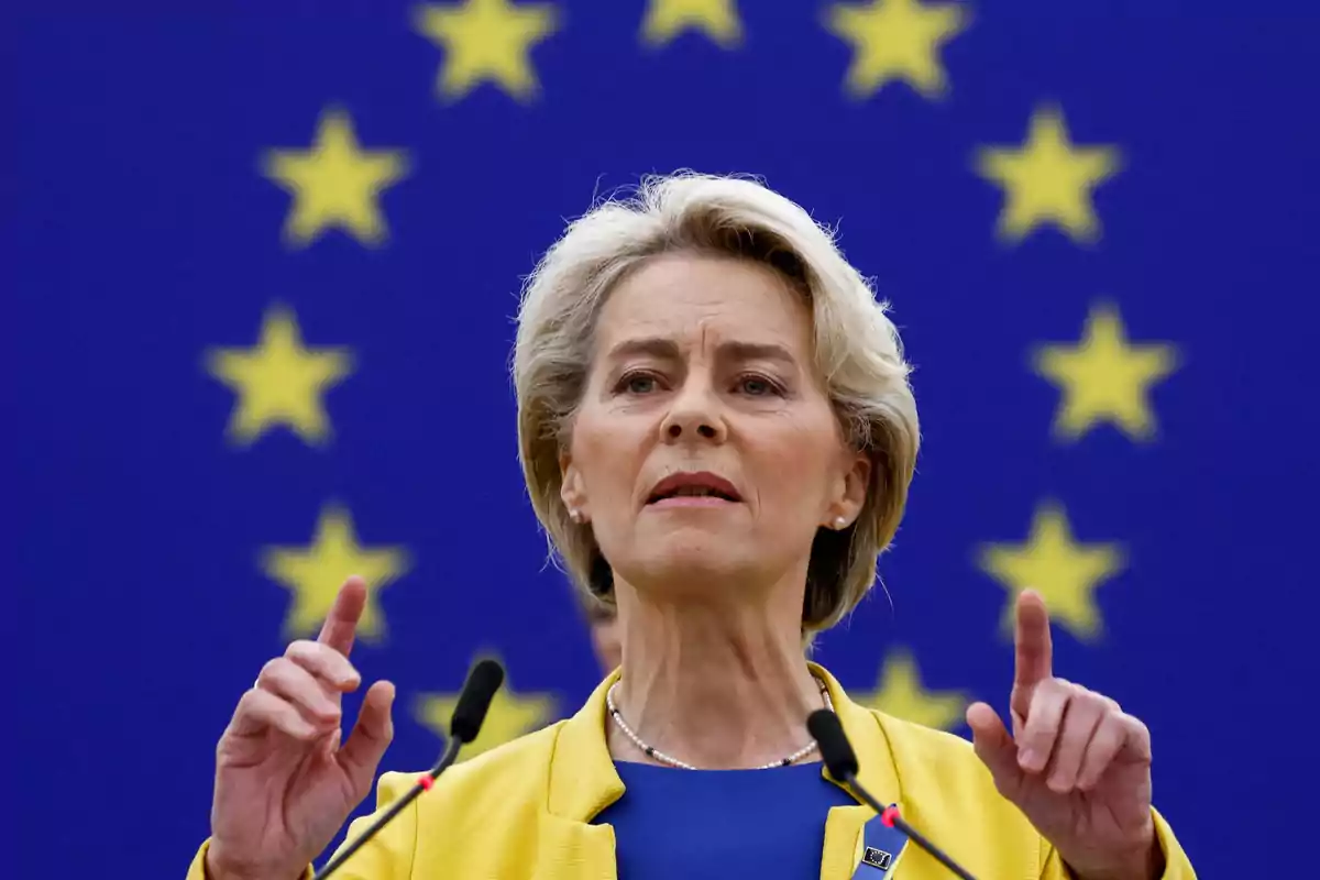 Mujer de cabello rubio hablando frente a micrófonos con fondo de bandera de la Unión Europea