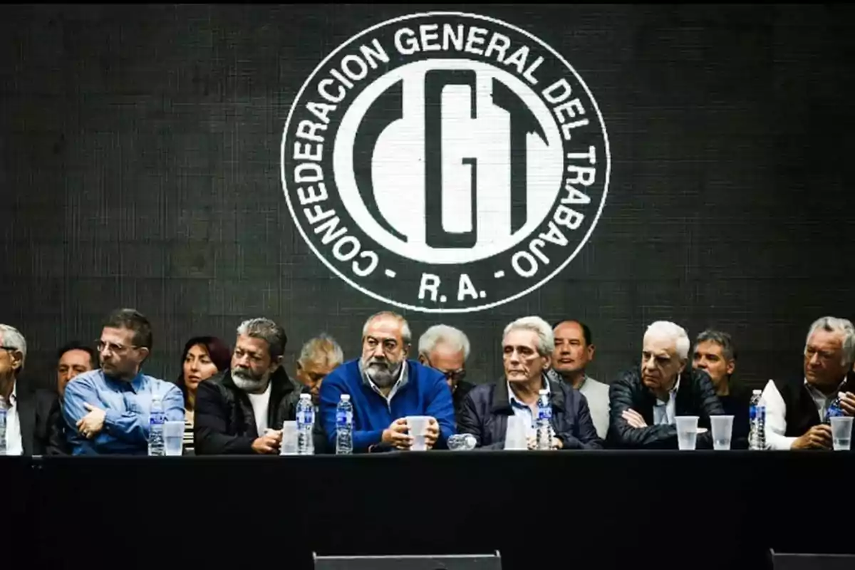 A group of people sitting at a table in front of a large logo of the General Confederation of Labor of the Argentine Republic