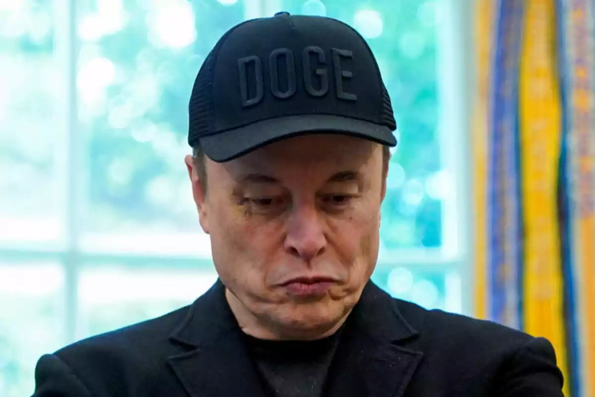 Hombre con gorra negra que tiene la palabra DOGE mirando hacia abajo con expresión seria