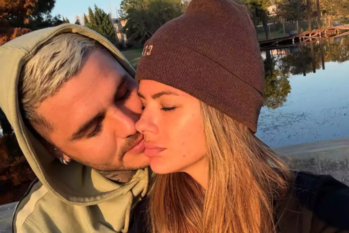 Una pareja se muestra abrazada al aire libre junto a un lago mientras el hombre besa tiernamente la mejilla de la mujer que tiene los ojos cerrados y lleva un gorro marrón.