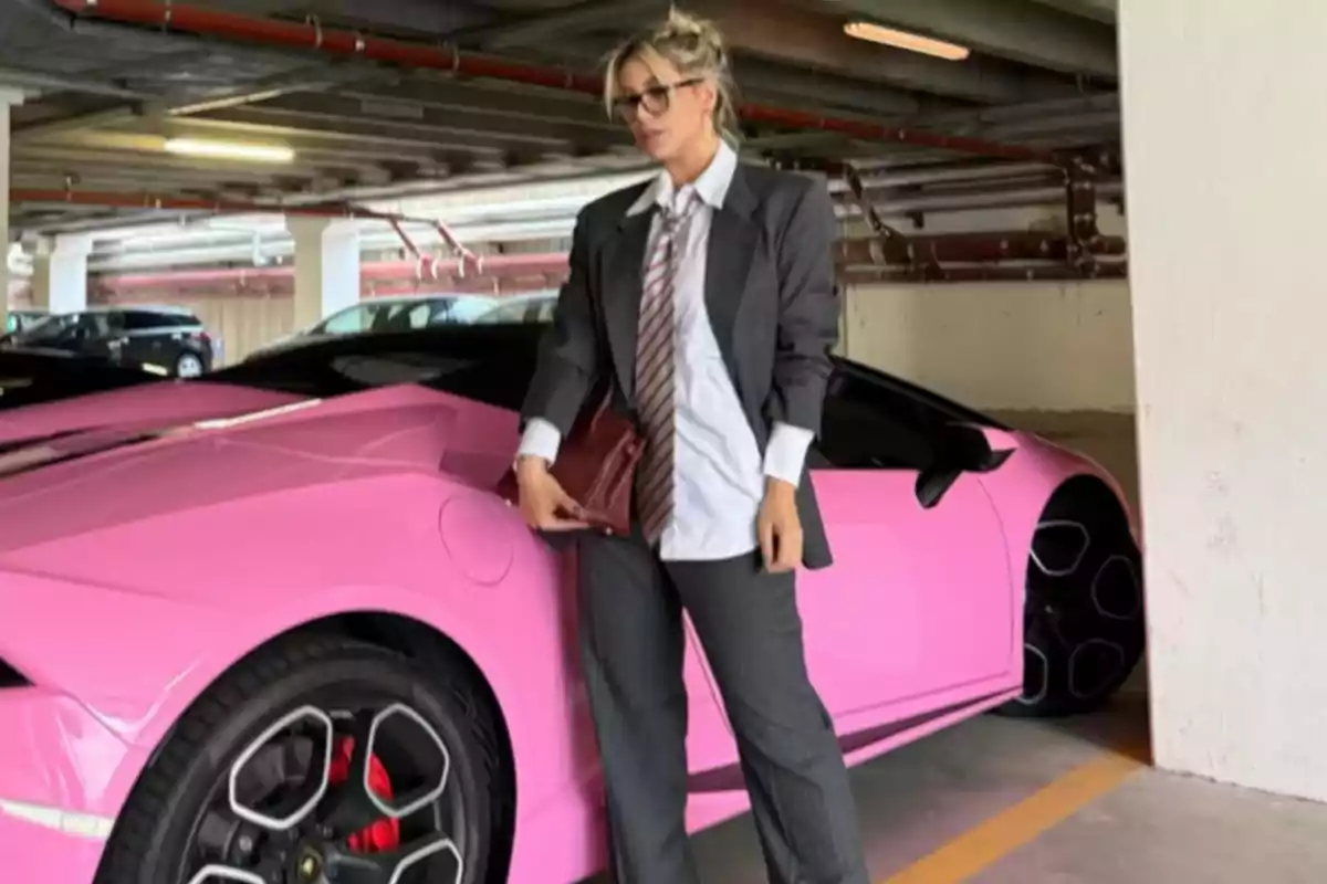 Mujer con traje gris y corbata posando junto a un auto deportivo rosa en un estacionamiento techado Mujer con traje gris y corbata posando junto a un auto deportivo rosa en un estacionamiento techado