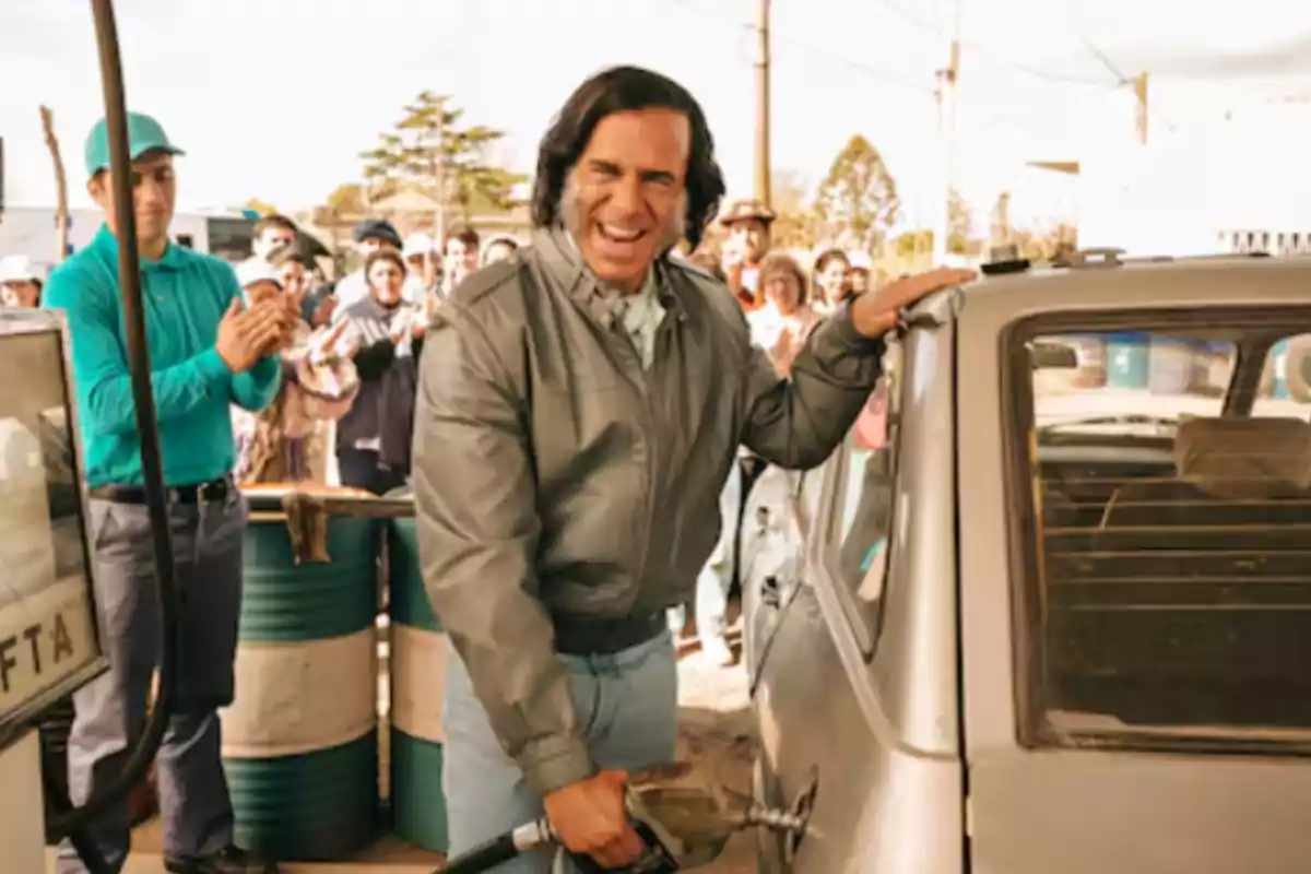 Smiling man pumping gas into a car while a crowd watches and applauds at a gas station
