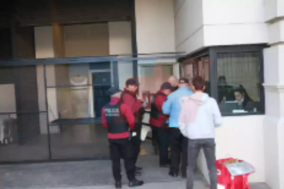Varias personas, incluyendo policías, se encuentran reunidas frente a una caseta de vigilancia en la entrada de un edificio.