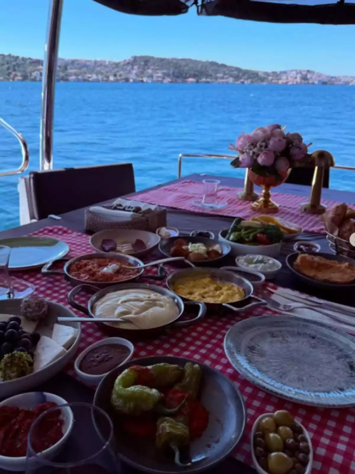 Table with a red and white checkered tablecloth, set with various dishes and appetizers, located on a boat overlooking the sea and hills in the background Table with a red and white checkered tablecloth, set with various dishes and appetizers, located on a boat overlooking the sea and hills in the background