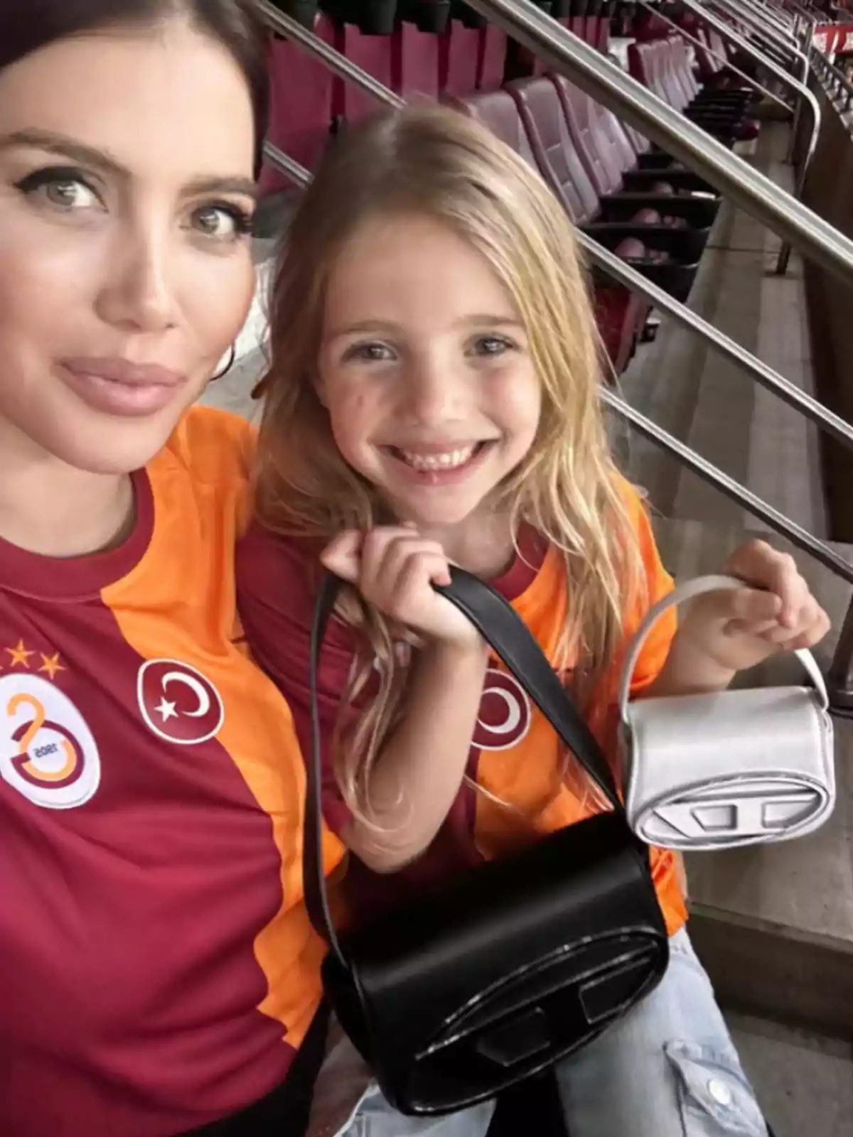 Mujer y niña sonriente con camisetas del Galatasaray posan en un estadio mientras muestran dos bolsos, uno negro y uno plateado. Mujer y niña sonriente con camisetas del Galatasaray posan en un estadio mientras muestran dos bolsos, uno negro y uno plateado.