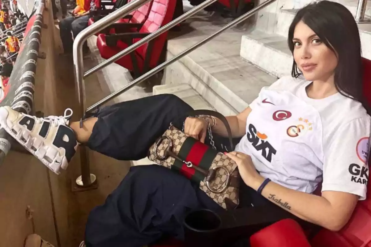 Mujer sentada en un estadio con camiseta blanca de fútbol, pantalones oscuros, tenis blancos y bolso de diseñador, mirando a la cámara mientras apoya los pies en la barandilla. Mujer sentada en un estadio con camiseta blanca de fútbol, pantalones oscuros, tenis blancos y bolso de diseñador, mirando a la cámara mientras apoya los pies en la barandilla.