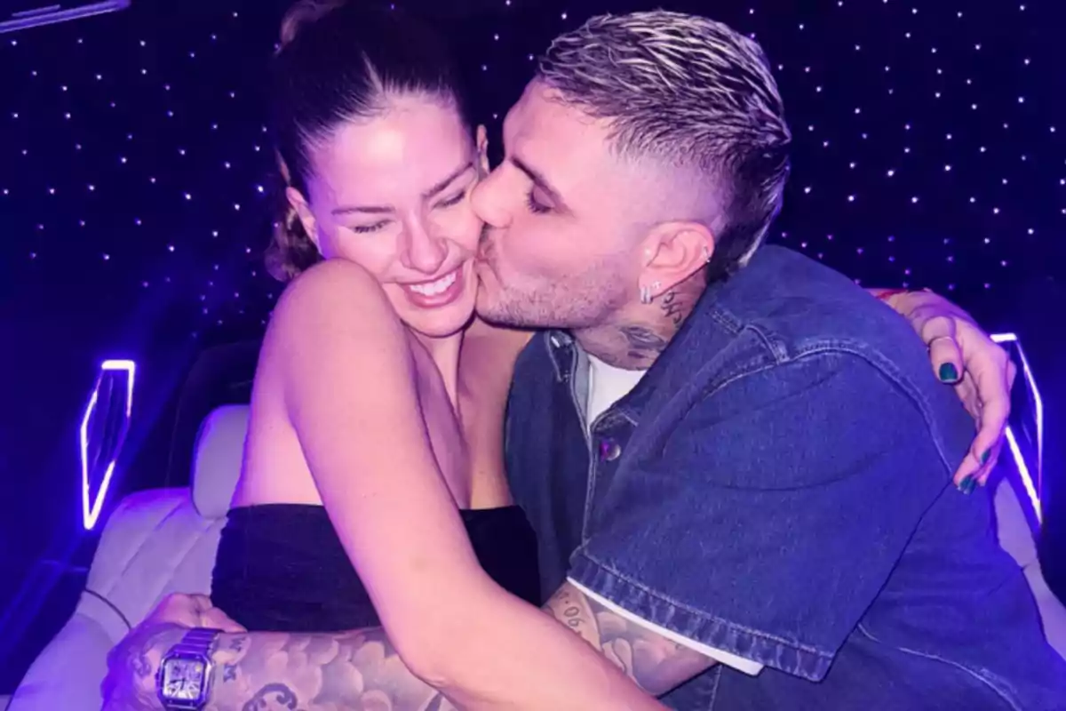 Pareja abrazándose y sonriendo mientras el hombre besa la mejilla de la mujer en un ambiente iluminado con luces moradas Pareja abrazándose y sonriendo mientras el hombre besa la mejilla de la mujer en un ambiente iluminado con luces moradas