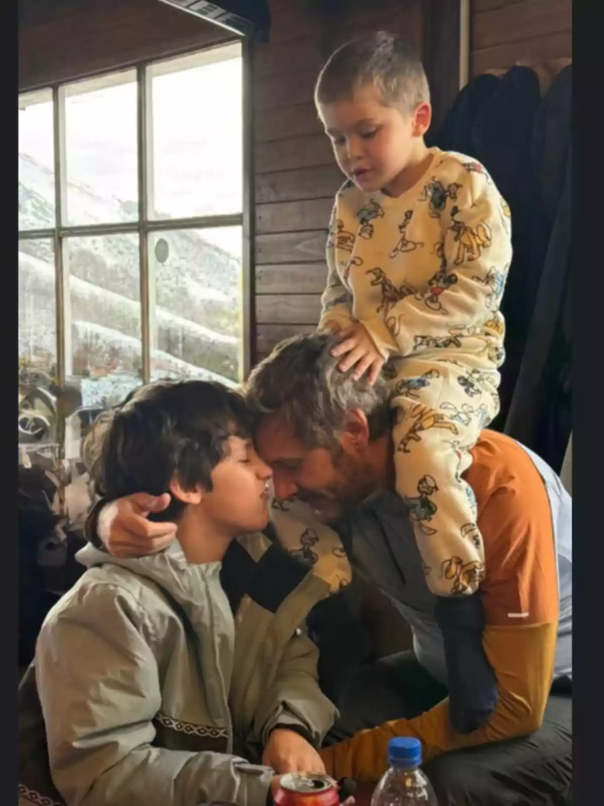 A man hugs two children inside a wooden cabin while one of the children sits on his shoulders and the other receives a kiss on the forehead. A man hugs two children inside a wooden cabin while one of the children sits on his shoulders and the other receives a kiss on the forehead.