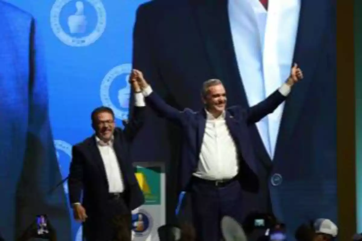 Dos hombres de traje levantan las manos en señal de victoria frente a una pantalla azul con logotipos circulares y un pulgar hacia arriba