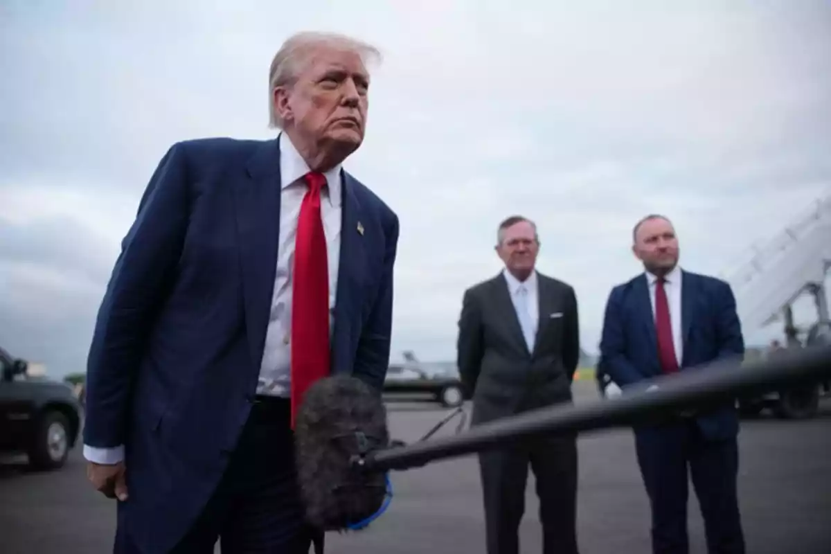 A man in a blue suit and red tie leans toward a microphone while two men in suits watch him in the background on an airport runway. A man in a blue suit and red tie leans toward a microphone while two men in suits watch him in the background on an airport runway.