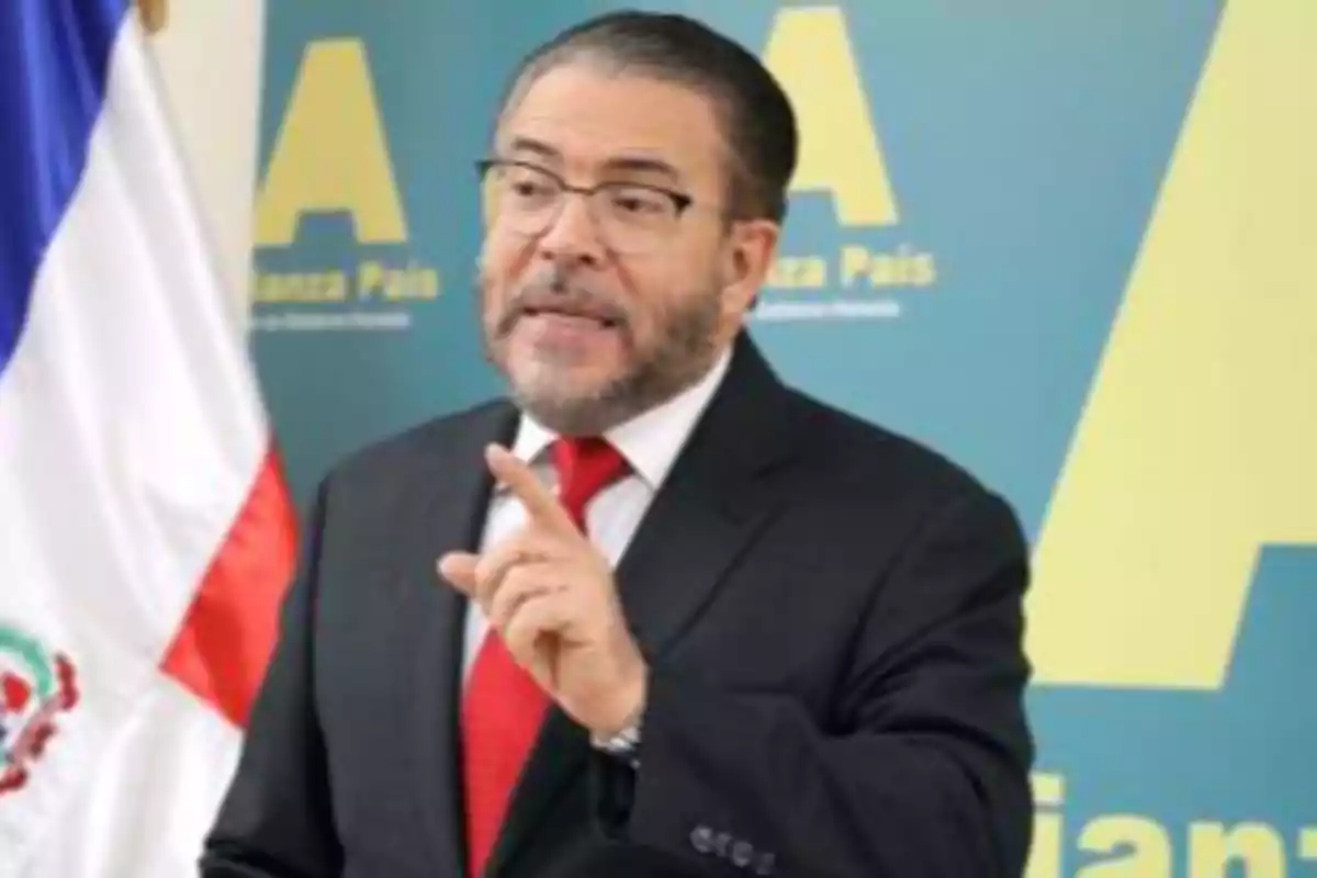 Hombre con gafas y barba vestido con traje oscuro y corbata roja hablando frente a un micrófono con una bandera y un fondo azul con letras amarillas