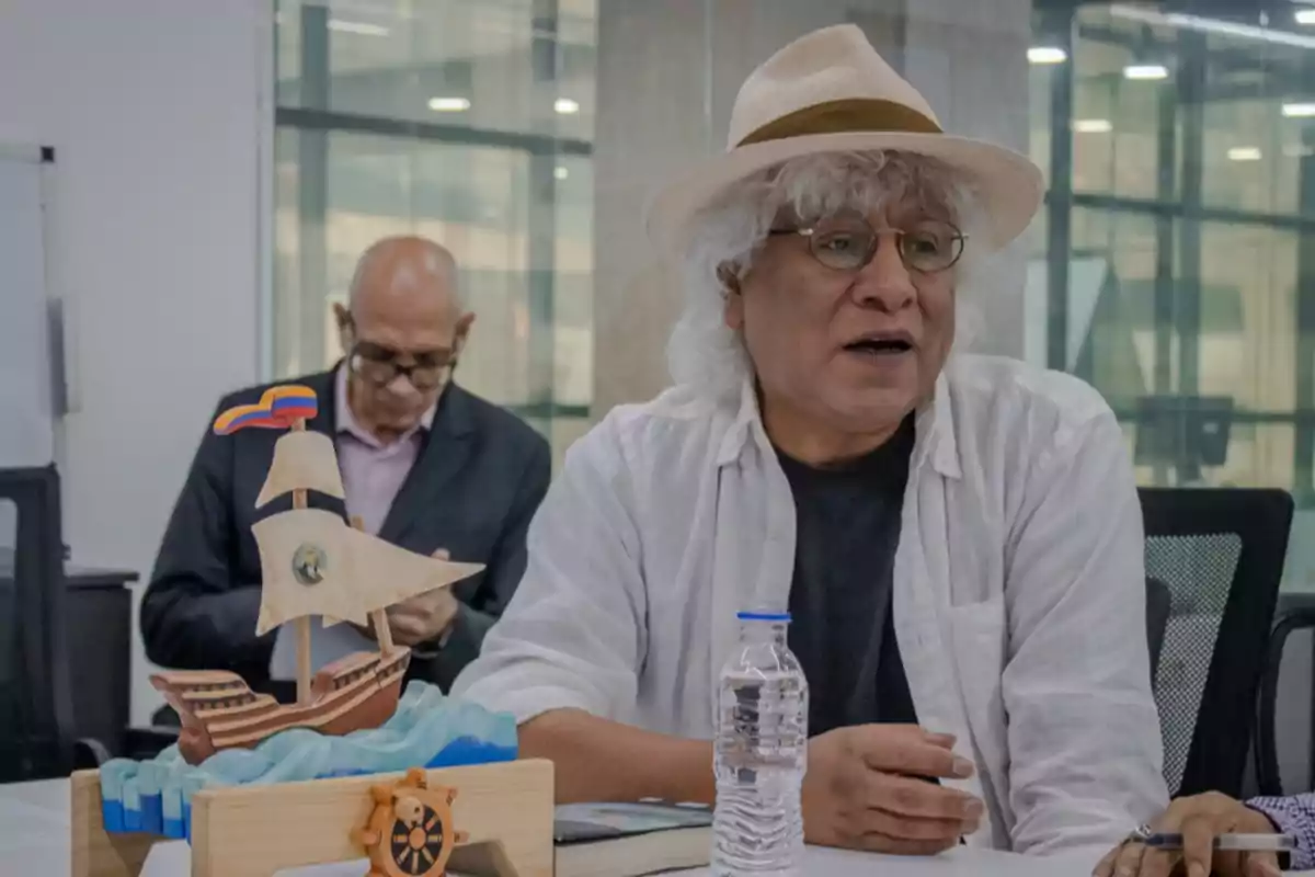 Hombre de cabello canoso y sombrero sentado en una mesa junto a una maqueta de barco y una botella de agua mientras otra persona está al fondo revisando papeles Hombre de cabello canoso y sombrero sentado en una mesa junto a una maqueta de barco y una botella de agua mientras otra persona está al fondo revisando papeles