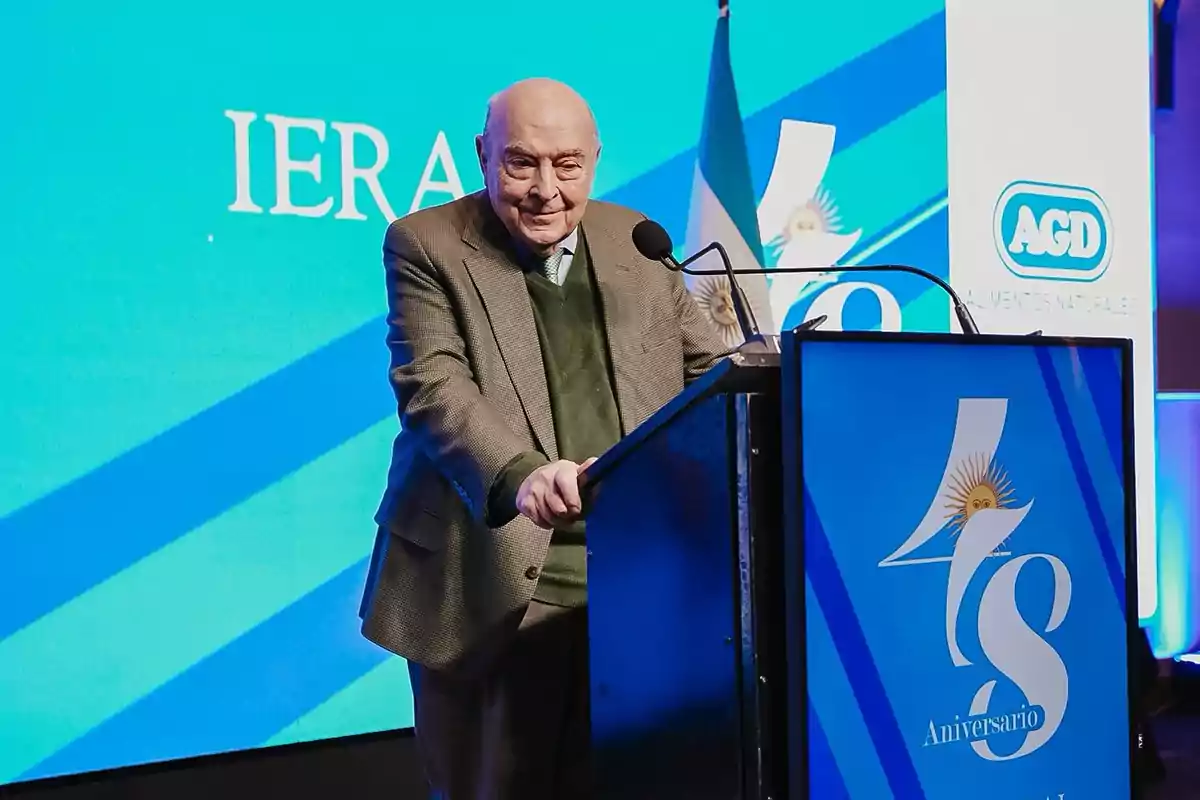 Hombre mayor de pie detrás de un atril durante un evento de aniversario con fondo azul y bandera argentina